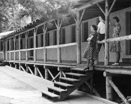 Teenage girls pose at the Beulah resort near Pueblo, Colorado, on the log railing of an open porch fronting a frame, one-story dormitory. The walks at the bottom of the steps are concrete with curved 'grout' lines.