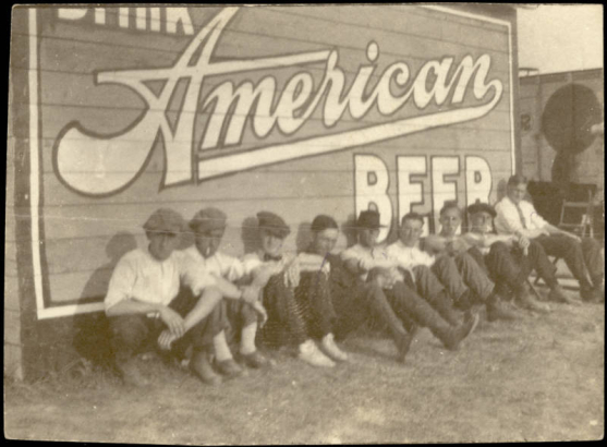 Young men, probably Devlin's Zouaves with the Sells Floto Circus and Buffalo Bill's Wild West Show, pose seated on the ground in Great Falls (Cascade County), Montana. The young men are near a billboard that reads: "Drink American Beer." The boys wear caps, shirts and pants. A circus trailer is seen in the background.