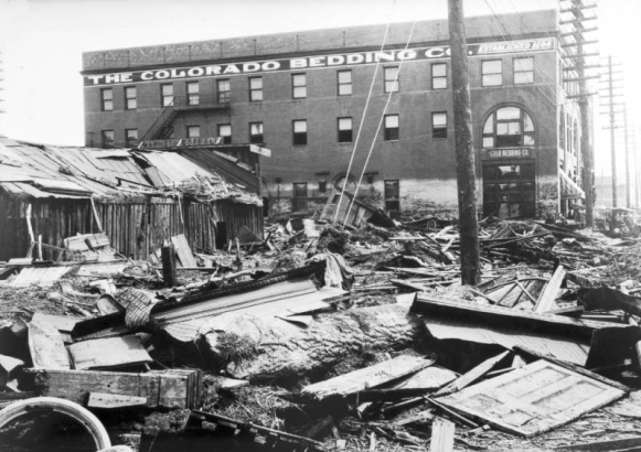 Flood debris in Pueblo, Colorado, includes carved wood molding, stamped ceiling metal, door, crates, wheels, lumber, and mud. A frame building has roof damage and a sign: "Mammoth Corral." A shed lays sideways in front of a four-story brick warehouse with arched windows and signs: "The Colorado Bedding Co. Established 1894." White residue covers the first  floor, showing the peak of the high water.