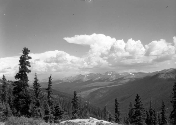 Pine trees stand on a hillside in the foreground in This view looking north toward a ridge of mountains in the distance from Berthoud Pass on the border of Clear Creek and Grand counties in Colorado. Small patches of snow are on the mountains in the distance. Cumulus clouds form a thunderhead anvil in the distance.