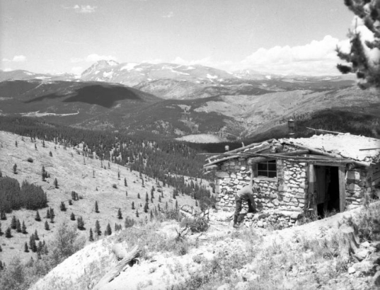 An unidentified man leans over, possibly to tie his shoe, in front of stone cabin in the foreground in This view near Jenny Lind Gulch, a tributary of South Boulder Creek near Apex in Gilpin County, Colorado. Pine trees line the rolling hills leading back to a ridge of mountains in the distance.