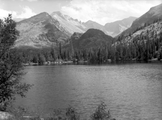 Rugged mountains stand in the distance in This view of Bear Lake in Rocky Mountain National Park in Larimer County, Colorado. Tall pine trees stand on the far shore of the lake.