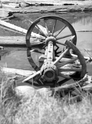 A set of wagon wheels sits half submerged in the water on the edge of Lost Lake west of Eldora in Boulder County, Colorado. A portion of the nearer wheel is missing. Pieces of wood float near the set of wheels.
