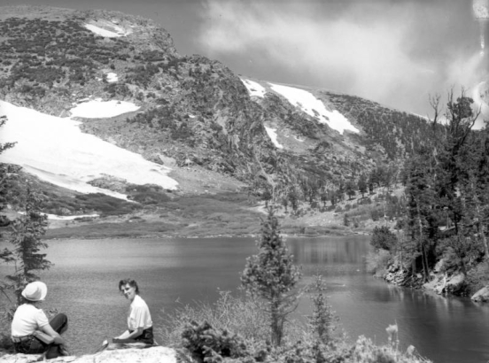 Two unidentified women sit on rocks in the left foreground on the  near shore of St. Mary's Lake looking toward St. Mary's Glacier in Clear Creek County, Colorado. Tall trees stand on the shore of the lake in the right midground and near where the two women sit.