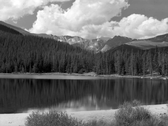 View of Echo Lake near Mount Evans in Clear Creek County, Colorado; pine forest and narrow beaches surround the water. Cumulus clouds top snow dotted mountain peaks in the distance.