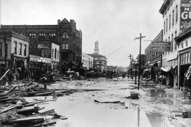 Water, mud and debris fill Union Avenue in Pueblo, Colorado. Brick storefronts show signs: "Klein & Leiser," "Colonial Jewelry," "Exchange Rooming House," "Weisel Seed," "Star House," and "Economy Clothing." Cast iron street lamps with milk glass globes line the thoroughfare. The Board of Trade Building, City Hall, and the turret of the Riverside Block show above other structures. The steel arch of the Arkansas River bridge is past wrecked automobiles and piled lumber.