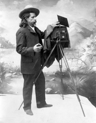 Photographer J. B. Sturtevant, nicknamed "Rocky Mountain Joe," stands near a large camera tripod in front of a studio backdrop painted with a landscape scene. Sturtevant, born in Boston, Massachusetts, became the first regular photographer in Boulder.Sturtevant wears a suit, a moustache, and a small pointed beard; he holds a lens in his right hand and a square plate in his left hand.  A cloth hangs over the back of the camera.