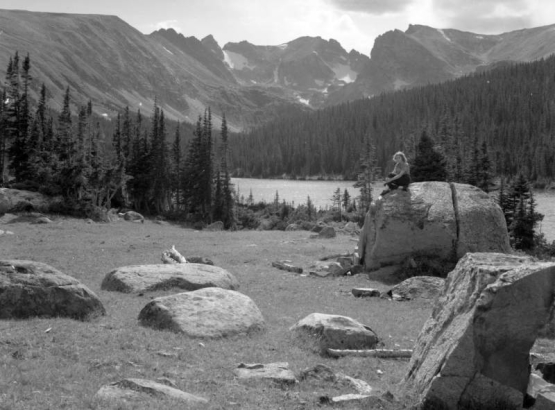 An unidentified woman sits on a large boulder near Long Lake west  of Ward in Boulder County, Colorado. Additional smaller boulders are also in the foreground. Tall pine trees stand along the shore of the lake in the  background. A ridge of mountains is in the distance.