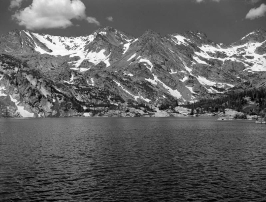 Silver Lake stands in the foreground in Boulder County, Colorado,  with a ridge of mountains in the distance. Tall pine trees stand on the far shore of the lake. Patches of snow are on the distant peaks. Large clouds are in the sky.