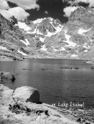 A large rock and tree branches stand in the foreground on the shore of Lake Isabelle in Boulder County, Colorado. Several smaller rocks also lie in the foreground. A ridge of jagged mountains with patches of snow on them is in the distance on the far shore of the lake.