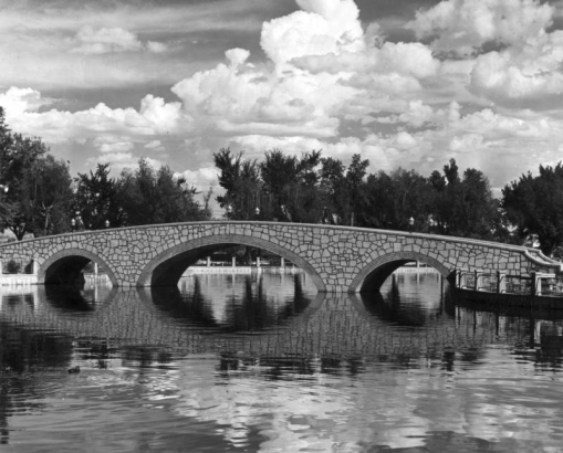 Arching bridge over Lake Clara at Mineral Palace Park in Pueblo, Colorado, is of random, pale stone with dark grout. Arches underneath provide support, and all is reflected in the rippled water in the foreground.
