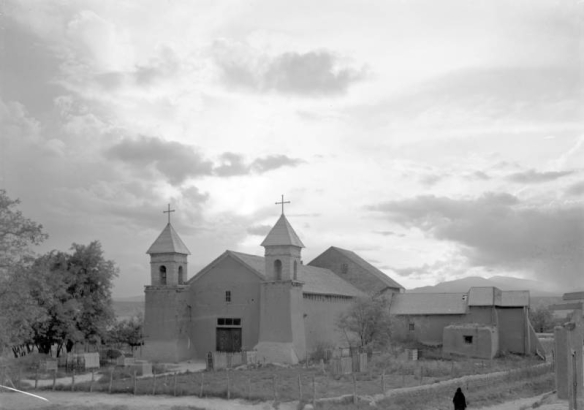 The Spanish Colonial mission at Santa Cruz, New Mexico is adobe and has two pitched-roof steeples with crosses on top. A woman with a dark shawl wrapped over her head and shoulders walks in the foreground down a path at the side of the mission.