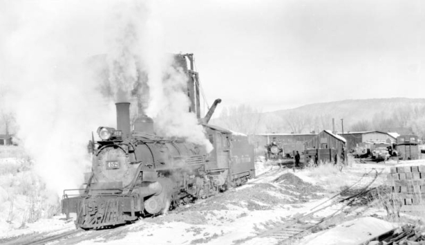 Three-quarter view of left side of engine, from front end, close view; at water tank; smoke, steam, snow; shops in background. Photographed:  Ridgway, Colorado, November 18, 1951.