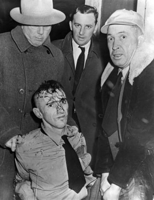 Werner Carl Schwartzmiller, recaptured after leading the break from the State Penitentiary in Canon City, Colorado, glares as Warden Roy Best pulls his shirt. Blood on his head, face and clothes is the result of hammer blows dealt by Mrs. Oliver, whose family he held captive for several hours. He wears a tie; chains and cable bind his arms in back. Wann and Whitelow look on, the latter wears a fur collared coat and baseball cap.