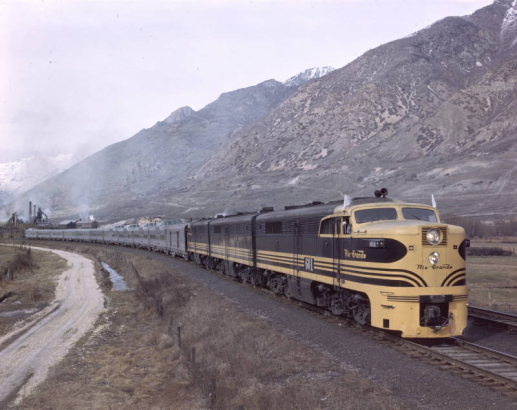 View of the eastbound California Zephyr, pulled by Denver & Rio Grande Western locomotive #601, in the Salt Lake Valley, Salt Lake County, Utah. Shows the train, with domed observation cars, near the Wasatch Mountains. The KF Steel plant is in the distance.