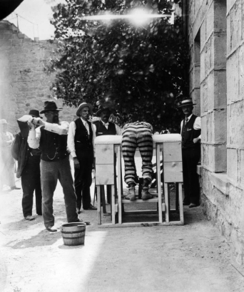 Bent over a wooden, upholstered frame, a prisoner at the State Penitentiary in Canon City, Colorado, wears patched, striped prison pants. His ankles are shackled, and a man in a vest, gold chain and cowboy hat swings a hinged paddle, targeting the convict's buttocks. A wooden bucket is on the ground, for wetting the implement. Men in vests, ties, and straw hats look on.