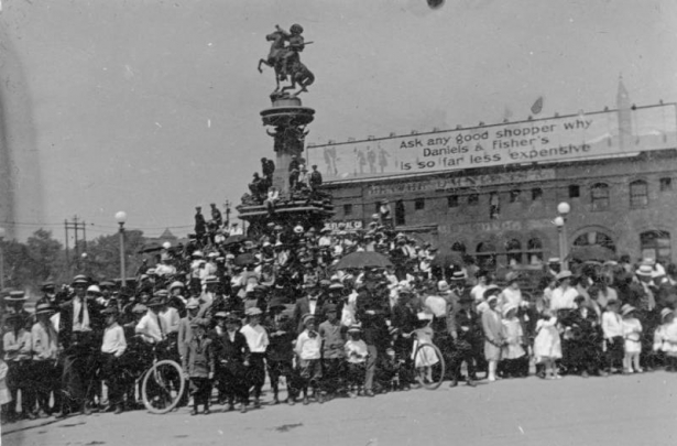 People crowd around the Pioneer Monument during a parade for the Sells Floto Circus and Buffalo Bill's Wild West Show in Denver, Colorado; a billboard reads: "Ask Any Good Shopper Why Daniels and Fisher is so Far Less Expensive."