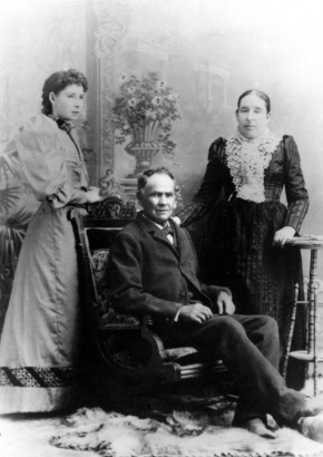 Studio portrait of Magdalena Córdova, daughter of Juan Bautista Córdova, and his wife, María Quirina Sánches. Magdalena wears a long dress with leg of mutton sleeves, and velvet trim. Her hair is curled and pulled back. She stands beside her father who is seated in a chair; he  wears a suit and bow tie, his hair is receding and he is clean shaven. His wife, María wears a dark silk dress with slightly puffed sleeves and a crocheted bertha collar.