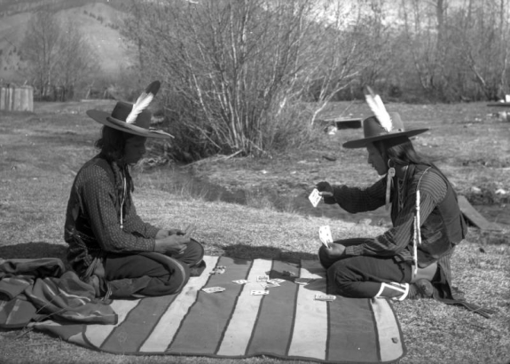 Antoine Moiese ("Grizzly Door") and Michael, two Native American men on the Flathead Indian Reservation in western Montana, sit on a striped blanket playing cards. Both men wear wide-brimmed hats with single feathers stuck in their bands. Both also wear vests and beaded necklaces. The man on the right has several ermine fur tassles hanging from his vest. A small creek is in the background with several shrubs along its bank.