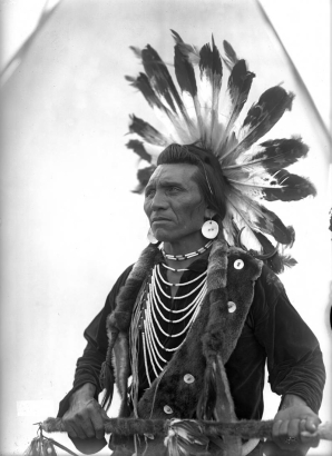Chief Eagle, a Native American man on the Flathead Indian Reservation in western Montana, poses wearing a warrior's headdress in front of a teepee on the reservation. He wears large, round earrings, numerous beaded necklaces hanging around his neck, an otter sash across his chest, and holds a long spear, possibly a coup-stick, also wrapped in fur.