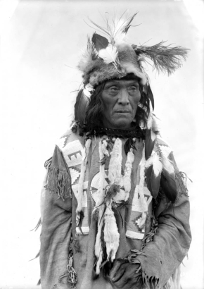 Francois Dead Horse, a Native American man on the Flathead Indian  Reservation in western Montana, poses in front of a teepee on the reservation. He wears a fur hat adorned with feathers, a buckskin shirt with beads sewn on it, and ermine fur tassles adorning it.