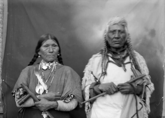 Studio portrait of Grizzly Bear and Marie, two Native Americans (Salish) on the Flathead Indian Reservation in western Montana. They pose in front of a painted canvas drop. Grizzly Bear holds a ceremonial pipe and wears a woven fringed tunic and has his long hair in braids, he has a scarf tied around his neck. Marie wears a polka-dotted dress and also holds a ceremonial pipe. She has several necklaces with an eagle feather hanging around her neck. Her hair is in two thick braids.