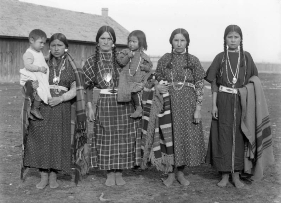 Four Native American women, including Louise Kumkumpoo Qumqmtpu, Agate Ogden Finley, Mrs. Latati and Agnes Incashola, pose with two small children on the Flathead Indian Reservation in western Montana. The women all wear their hair in braids. Their dresses are made from floral and checkered patterned materials. Each of them also wears several necklaces around her neck. Three of the women hold patterned, wool blankets. A long, wood building is in the left background.