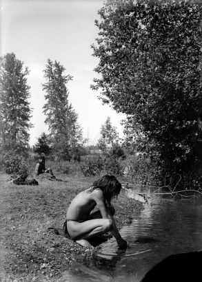 Antoine Ninamee and Loma Charlemain, two Native American boys on the Flathead Indian Reservation in western Montana, sit near a creek. The young man in the foreground wears a loincloth and squats down next to the creek scooping water into his hands. The other young man sits in the left background near a pile of clothing. He wears his hair in braids. Numerous trees stand along the creek.