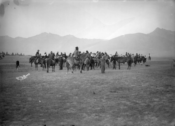 A group of unidentified Native Americans on horseback is gathered on the Flathead Indian Reservation in western Montana, possibly preparing for a twilight race. Many of the men wear wide-brimmed hats. Several men standing on the ground hold patterned blankets around their shoulders. A ridge of mountains is in the distance.