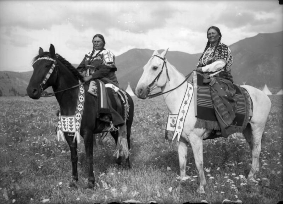 Two Native American women on the Flathead Indian Reservation in western Montana pose on horseback in a field. Both have long hair in braids; each wears a dress adorned with beads. The bridles on the horses are similarly adorned with beadwork. The women sit on patterned, wool blankets that cover their wood-frame saddles. Mountains aren the distance.
