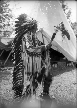 Joe Lamoose, a Native American on the Flathead Indian Reservation in western Montana, dances in front of a teepee on the reservation. He holds a pipe tomahawk adorned with fur and feathers and wears a long, feather headdress. Long, ermine fur tassles and beadwork adorn the sleeves of his buckskin shirt. He also wears beaded moccasins.