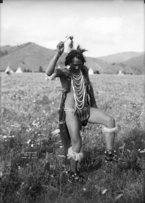 Joe Vanderburg (also knwn as Yellow Rock), a Native American man on the Flathead Indian Reservation in western Montana, dances in a field on the reservation. He wears a striped loincloth and several long, beaded necklaces. Strips of fur and bells are tied around his lower legs and his arms. Additional straps of fur are wrapped around his long braids. Several teepees are in the distance at the base of a ridge of mountains.