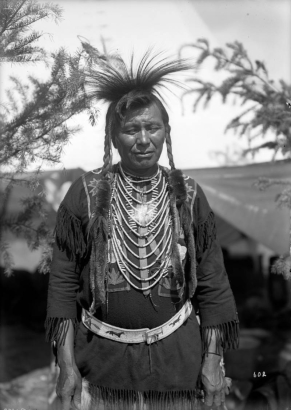 Loma Standing Bear, a Native American man on the Flathead Indian Reservation in western Montana, poses for an informal, half-length portrait. He wears his long hair in braids with strips of fur wrapped around their ends. He also wears a breastplate, a belt with beadwork depicting horses, a beaded-flower shirt, and tassels on the sleeves and bottom of his shirt.