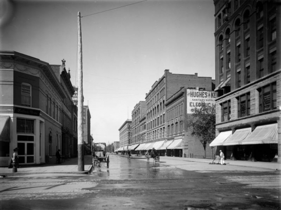 A man cleans the street in the right mid-ground at the intersection of California and 17th (Seventeenth) Streets in downtown Denver, Colorado. Businesses include the Colorado Cycle Supply Company, Cordes & Feldhauser Carpet Company, the Nathan Brothers, offices of Drs. J.W. Harris and J.B. Brown, and a restaurant. Signs advertise furnished rooms and "Mrs. Marrow's Home-Cooked Foods." Pedestrians and wagons travel on the dirt street.
