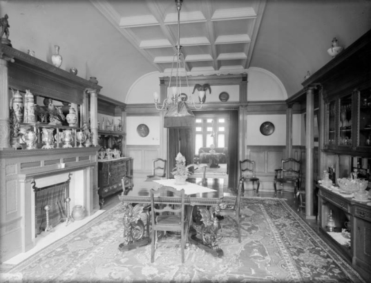 Interior view of a dining room in Denver, Colorado. Decor includes a table carved griffin legs, an Oriental rug, silver urns and vases, a stuffed bird, a crystal punch bowl, and a sideboard with a silver serving set. A ram's head is mounted above the door.