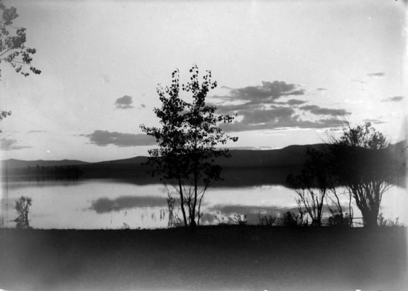 A few trees are on the near shore of Flathead Lake in western Montana in a view during sunset. Several clouds line the sky above a ridge of mountains in the distance.