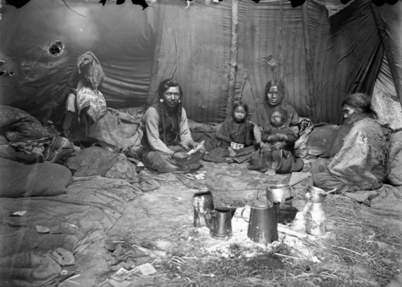 A group of unidentified Native Americans sits playing cards inside a teepee on the Flathead Indian Reservation in western Montana. Numerous pillows and blankets are on the ground inside the teepee. A group of pots and containers is in the foreground.
