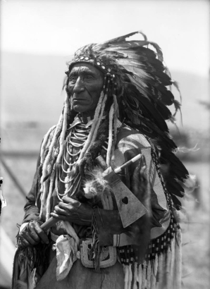 Joe Lamoose, a Native American man on the Flathead Indian Reservation in western Montana, poses for an informal, half-length portrait. He wears a feather headdress and holds a pipe tomahawk in his hands. He wears several beaded necklaces, ermine fur tassles and beads on the sleeves of his shirt, and a beaded pouch on his belt.