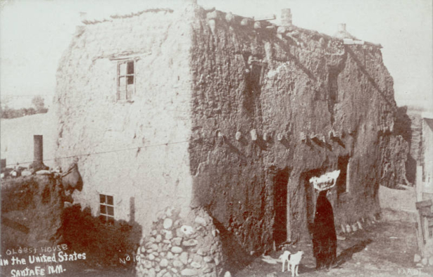 View of a dilapidated adobe house located at 215 E. De Vargas Street, Santa Fe (Santa Fe County), New Mexico. The house has vigas, chimneys and a stone buttress, and is thought to be the oldest house in the city. Shows a woman with a basket on her head and a dog.