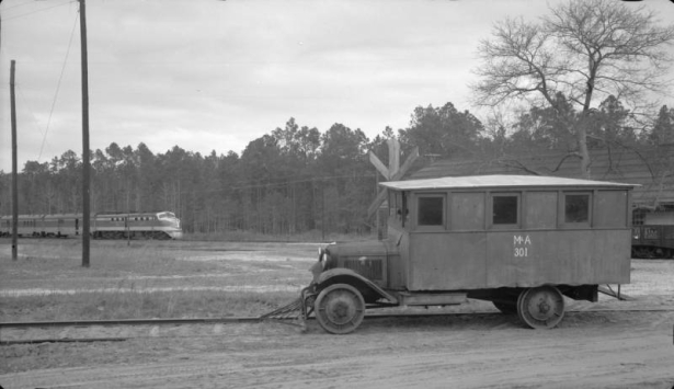 A Mississippi & Alabama railroad auto railer truck is parked on a siding near a road grade crossing in Vinegar Bend (Washington County), Alabama. The truck has a cowcatcher on the front. Lettering reads: "M&A 301." Shows the Gulf, Mobile and Ohio streamliner the "Rebel" in the distance with a diesel electric locomotive.