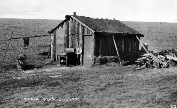 View of make-shift one-room cabin with sod roof, Gillett, Colorado. The small cabin is constructed of vertical logs, and a tar paper door and window. Two posts appear to support the gable roof. Materials around the cabin include: pile of scrap wood, wooden barrels, and a clothes  line connected to the house.