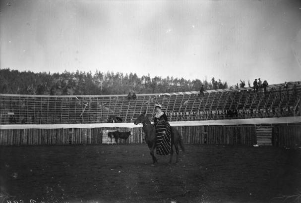 In This view of "La Cherita" participating in a bullfight in Gillett, Colorado, a woman in Mexican dress rides sidesaddle atop a horse. Wooden bleachers are in the background of the arena, with a few observers on the wooden bleachers. Bulls are in the background.