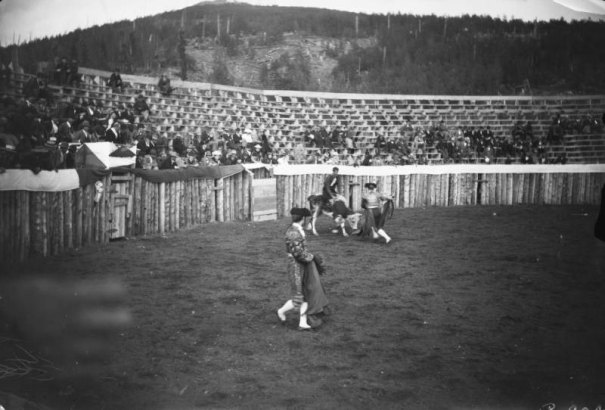 View of a bullfight in a small stadium, in Gillett, Colorado, show two men in bullfighting attire. Two matadors hold capes, a third sits atop a bull who is ready to charge. The bullfighters are in a wooden corral, while observers watch from bleachers. The matador's costumes include hat, embroidered bolero jacket, epaulets & pants.