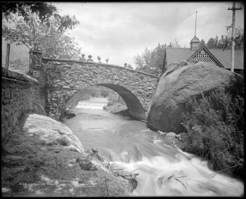 Rock footbridge crossing Fountain Creek to Soda Spring pavilion, Manitou Springs, Colorado; shows men and women standing on bridge.