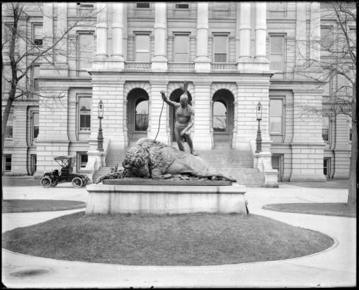 "Closing Era" bronze statue in front of Capitol building, Denver,  Colorado; made by Preston Powers, ca. 1890; presented to city in 1892 by women of Fortnightly Club. The statue is a lifelike size bronze Native American male standing over a slain buffalo. Front steps and second story of Capitol are in background; an early automobile is parked next to capitol; and electric lightposts appear on Capitol steps.