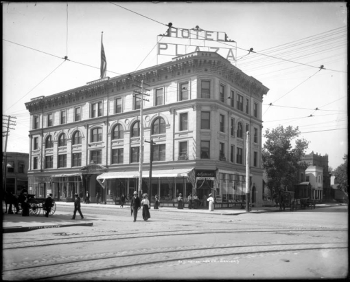 Exterior view of Plaza Hotel, 330 15th (Fifteenth) and corner of Tremont, Denver, Colorado, a four-story building with window awnings, Plaza hotel sign, and United States flag flying atop pole on roof; shows men and women on sidewalks and crossing street with street railway tracks, Manie Hyman's Cigar and Importing Company located in corner of building, and a delivery wagon, center right.