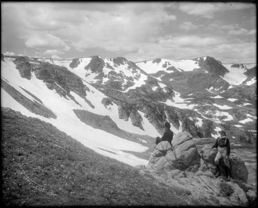 Looking to snow-covered Arapahoe Peaks, Colorado, from Denver and Salt Lake Railroad's Moffat Road; two men posed at boulders right foreground, one in suit and derby hat, and the other in knee-high laced boots, trousers tucked in boots, suspenders, long sleeved shirt with turtleneck and felt hat.