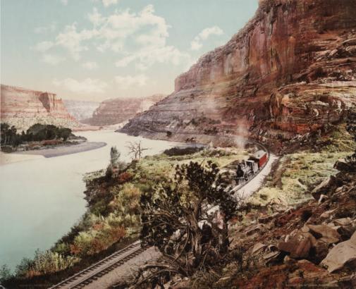 A Denver & Rio Grande railway train makes its way along the Colorado River in Ruby Canyon, in Utah. Sandstone cliffs and vegetation flank the water.
