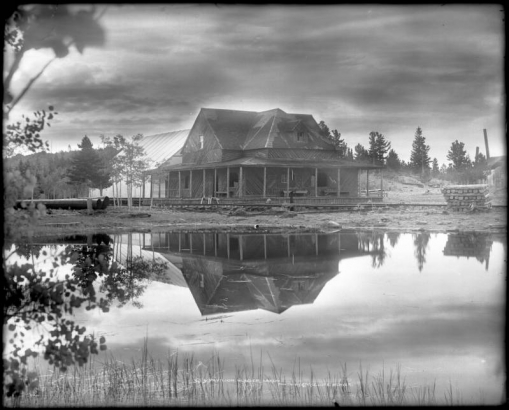 View of the edge of Glacier Lake and wooden frame picnic pavilion with canvas tent attached; shows the Colorado and Northwestern Railway narrow gauge rails either under repair or construction and the branch line to Eldora, Colorado. The building and dark clouds are reflected in the water.