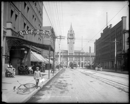 View down 17th (Seventeenth) street to Union Station prior to construction of the Welcome Arch; shows passengers boarding street railway car in center background, Oxford Hotel at left, Hendrie and Bolthoff Manufacturing and Supply Company at right, horse-drawn carriages on the street, and a young boy with knickers approaching bicycle left foreground; many men sit in front of the Oxford Hotel.